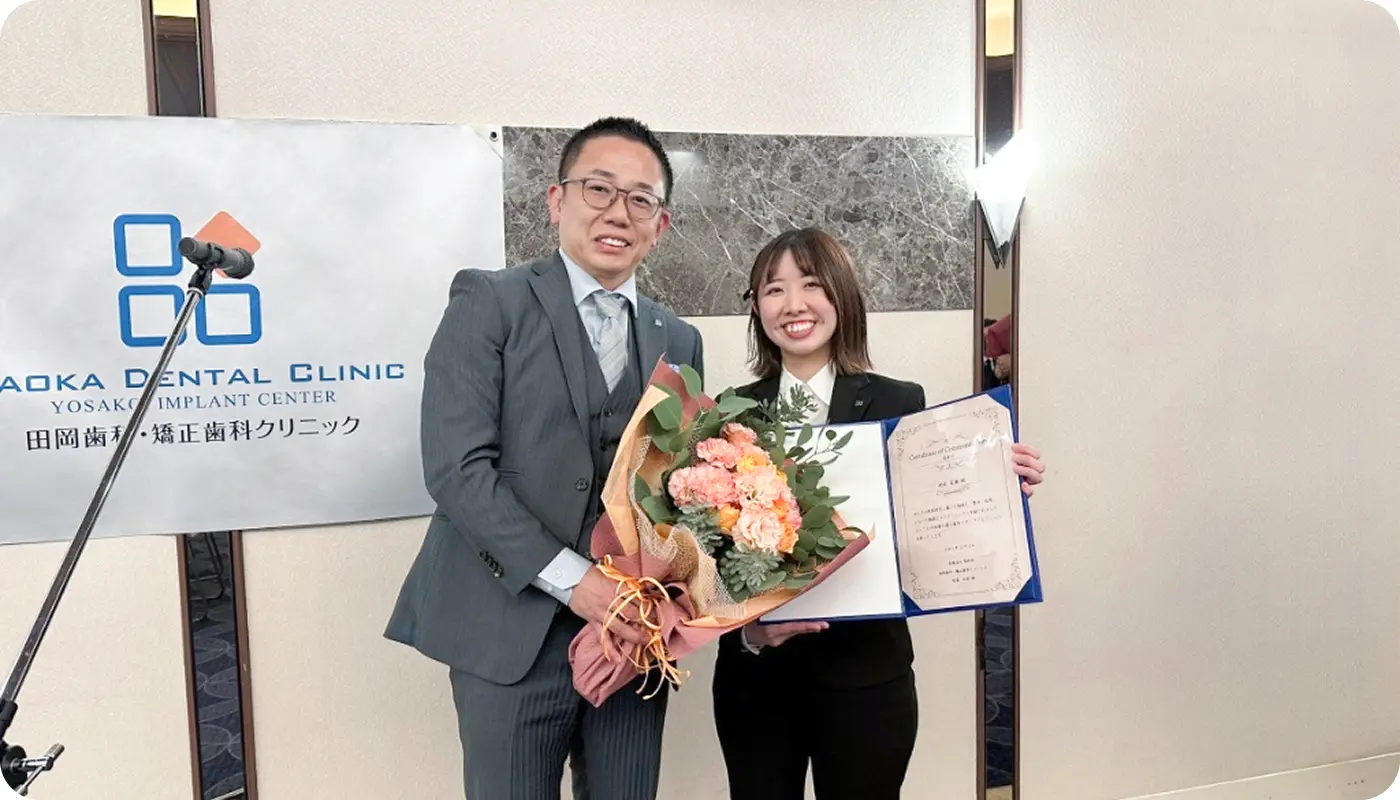 田岡歯科・矯正歯科クリニックの表彰式にて、院長から花束と表彰状を受け取り笑顔を見せる女性スタッフ。